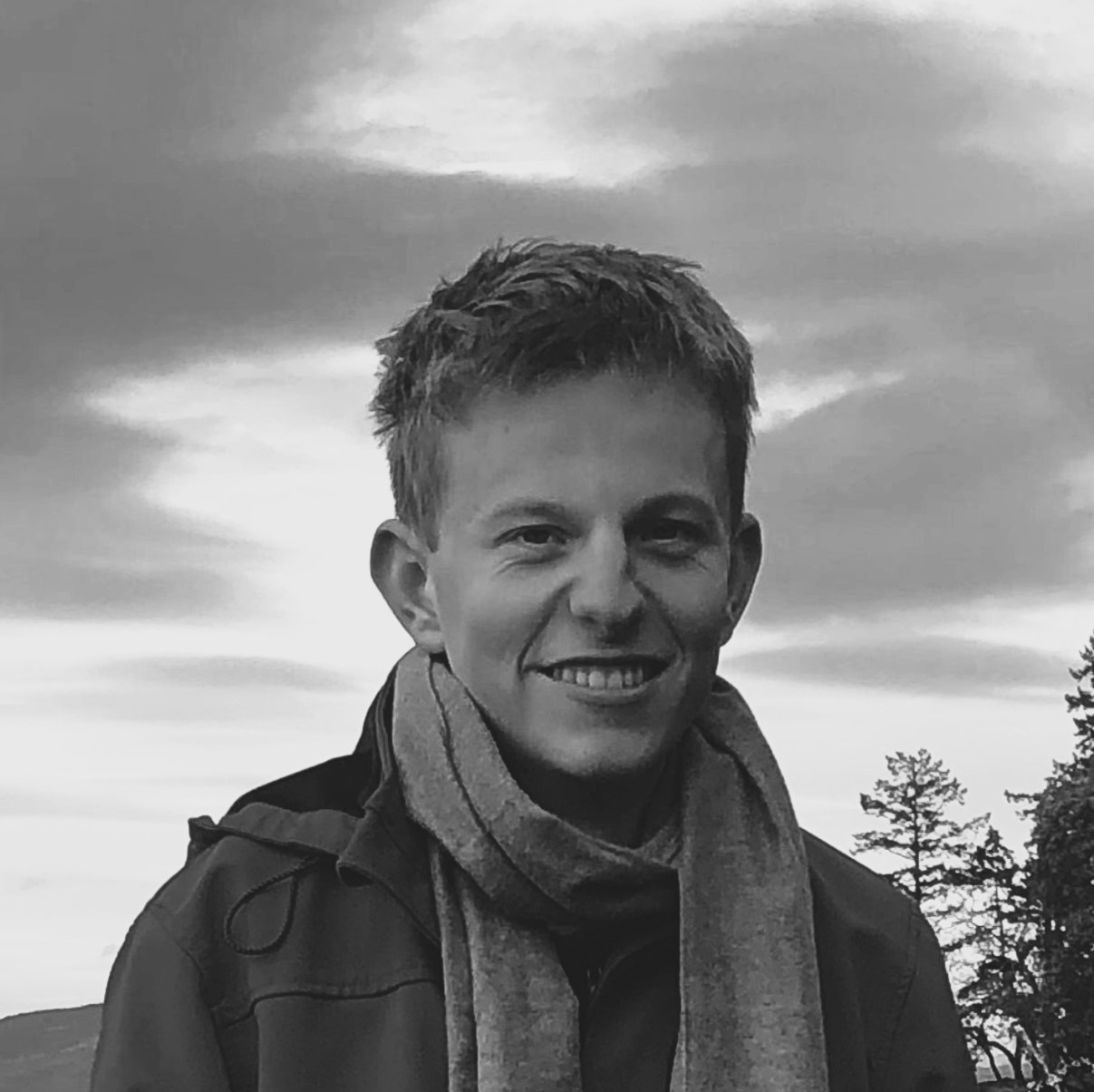 22 year old blonde man wearing a scarf and a rain jacket. He is outside and there are trees in the background. Black and white photo.